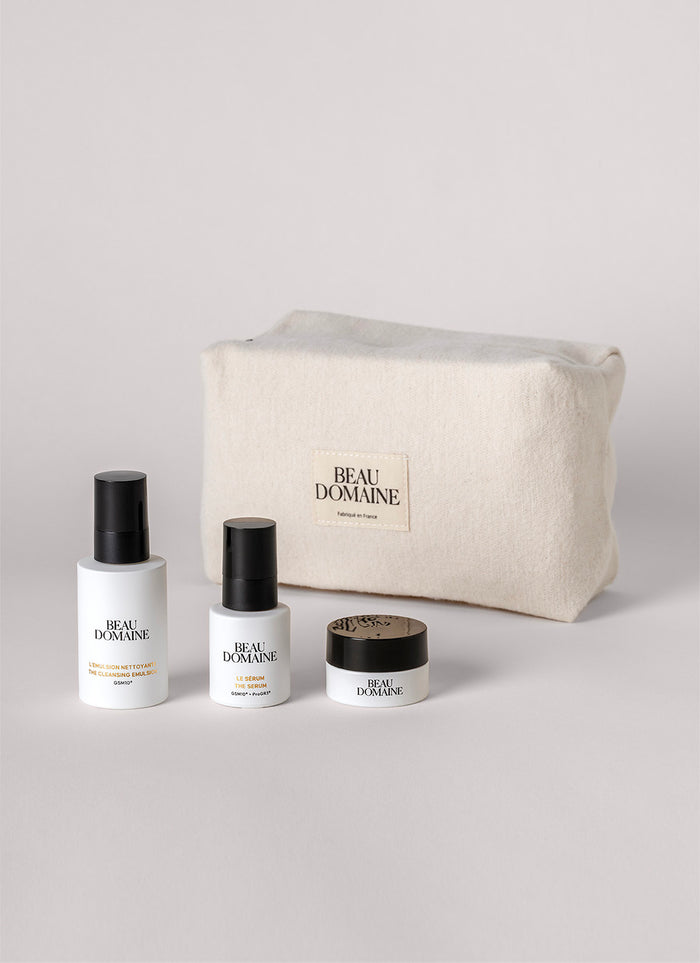 Set cosmetic cu sticluțe și un borcan în fața unei genți 'Beau Domaine' pe un fundal gri deschis